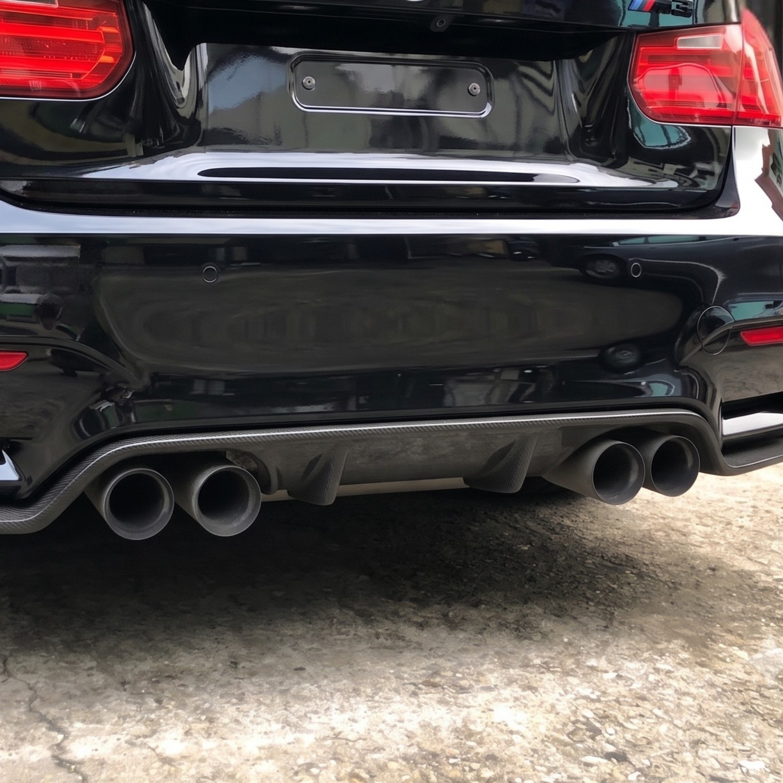 Rear view of a black BMW car showing the rear bumper carbon fiber diffuser with dual exhaust pipes, taillights, and the BMW roundel on the trunk.